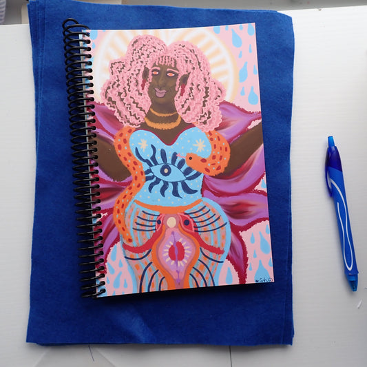 Notebook with a colorful illustration of a divine feminine goddess on a blue cover, placed on a white surface with a pen beside it.